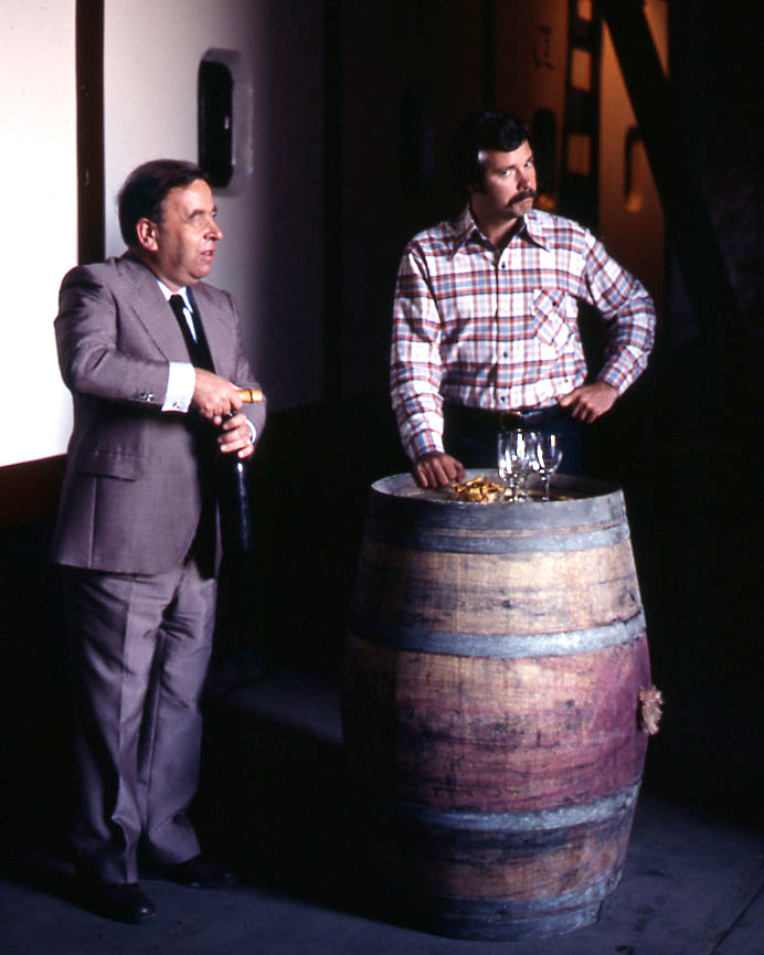A young John Sweazey on stage with another man, leaning against a wine barrel while the man opens a bottle of wine. Both men are well lit in the foreground, while the background is blurred and dark.