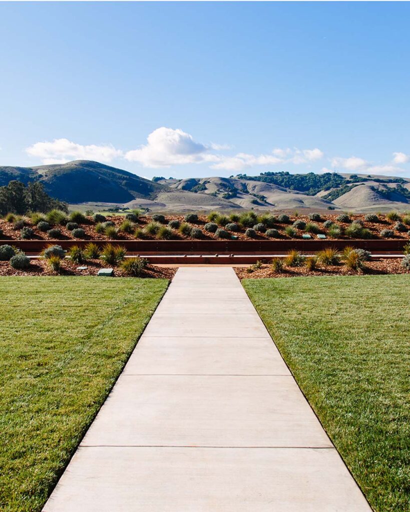 A long pathway in the centre of the foreground, leading to the bocce ball court, rolling mountains pictured in the background, under a cloudy blue sky.
