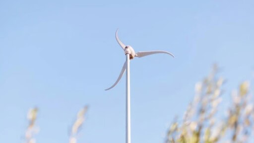 A Skystream wind turbine stands against a clear, light blue sky, with blurred foliage visible at the bottom of the frame.