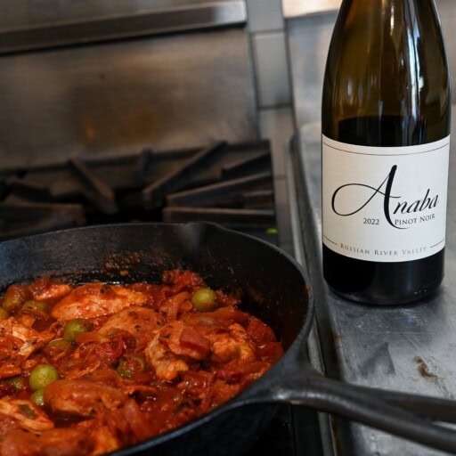 Braised chicken and olives in skillet with a bottle of Anaba Pinot Noir on counter next to it.